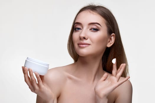 beautiful smiling girl applying skin care cream on her face and holding jar of cosmetic product on white background