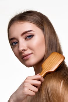 Beautiful smiling brunette woman looks at the camera and combs her hair with wooden brush isolated on white background