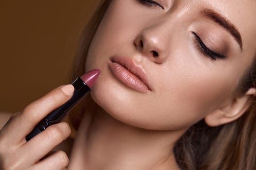 beautiful young smiling woman holding nude pink lipstick on beige background