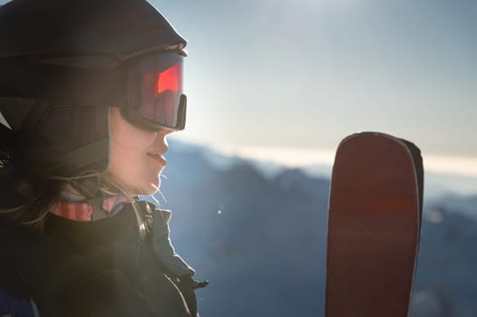 Portrait of sportswoman in helmet and mask with skis in hand, looking away, enjoying sunny frosty day, perfect day for skiing in ski resort.
