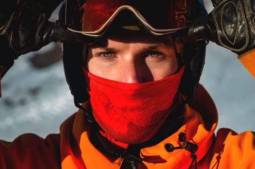 Portrait of a man with yellow eyes. Photo of a young serious sportsman in a helmet and ski goggles in his hands, against the backdrop of a snowy hill.