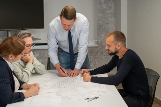 A caucasian man is standing making changes to a drawing, three colleagues are sitting at a table and listening to him