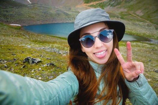 Selfie portrait of young attractive woman wearing sunglasses and backpack smiling with lake and mountains in the background, sunny morning.