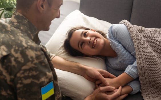 Soldier in Ukrainian military uniform kissing his daughter while she sleeping on sofa at home. Family reunion