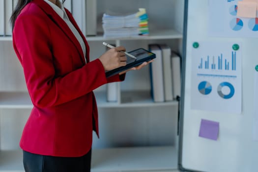 portrait of Beautiful young asian businesswoman presents business profits to colleagues at meeting, explaining business turn over on flipchart to coworkers in office with using tablet.