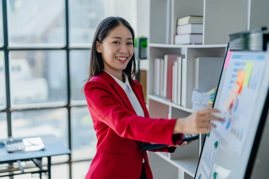 portrait of Beautiful young asian businesswoman presents business profits to colleagues at meeting, explaining business turn over on flipchart to coworkers in office with using tablet.