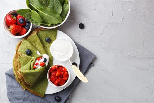 Green pancakes with spinach on a plate with fresh berries and sour cream, on a gray background with textiles. copy space