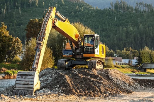 Yellow excavator (digger machine) digging stones, in river, forest in background