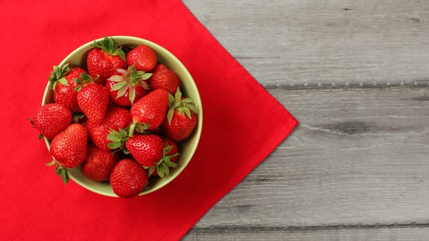 Red ripe strawberries in a small bowl, with tablecloth and gray desk under.