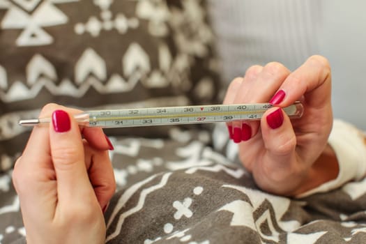 Detail on mercury thermometer held by hands of young woman laying in bed. Sick person checking body temperature.