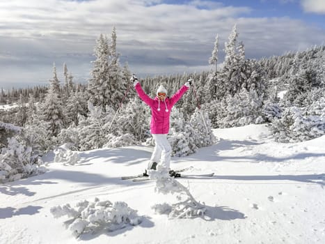 Young woman in pink jacket, boots, ski, poles, goggles and hat, hands in the air, looking happy, snow covered trees behind