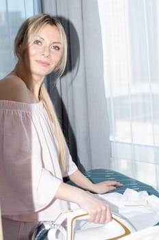 People. A beautiful Caucasian girl with long blond hair irons clothes on an ironing board against the background of a window, in pink home clothes with a bare shoulder. Concept.