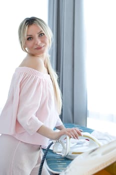 People. A beautiful Caucasian girl with long blond hair irons clothes on an ironing board against the background of a window, in pink home clothes with a bare shoulder. Concept.