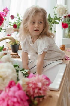 A little blonde girl on a kitchen countertop decorated with peonies. Spring atmosphere