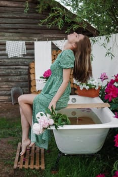 A woman is sitting on a cast-iron bathtub in the courtyard of a country house next to a bush of flowering peonies. The concept of summer, country life