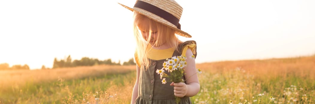 A little blonde girl in a straw hat walks in a field with a bouquet of daisies. The concept of walking in nature, freedom and an eco-friendly lifestyle