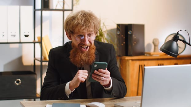 Oh my God Wow. Happy amazed young businessman guy working on smartphone shocked by sudden victory game winning lottery jackpot achievemen good news. Bearded redhead man freelancer at home office