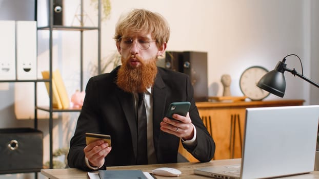 Bearded businessman programmer software developer working making online purchase payment shopping with credit bank card smartphone at modern home office desk with laptop. Young freelancer business man