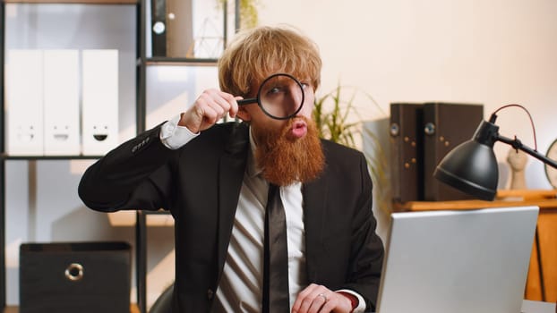 Investigator researcher scientist businessman working at home office holding magnifying glass near face looking into camera with big zoomed funny eye, searching, analysing. Professional freelancer man