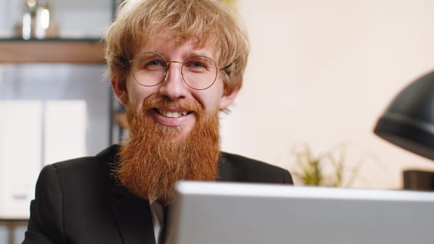 Close-up portrait young bearded businessman face freelancer at home office workplace working on laptop computer. Manager freelancer redhead man works on notebook send messages makes online purchases