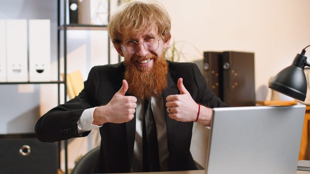 Like. Happy young bearded businessman guy working on laptop looking approvingly at camera showing thumbs up, like sign positive something good, positive feedback at office workplace. Freelancer man