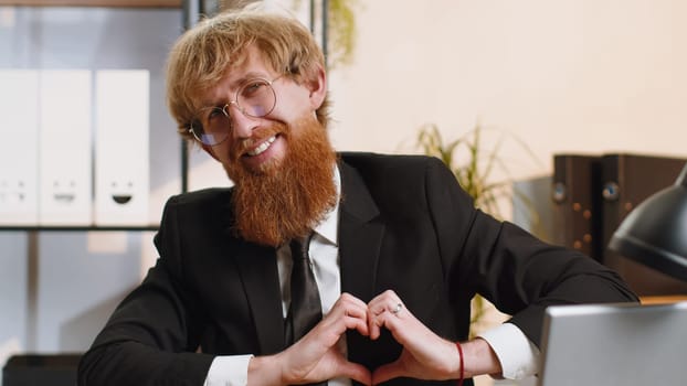 I love you. Young business man makes symbol of love, showing heart sign to camera, express romantic feelings, express sincere positive feelings at home office workplace. Charity, gratitude, donation