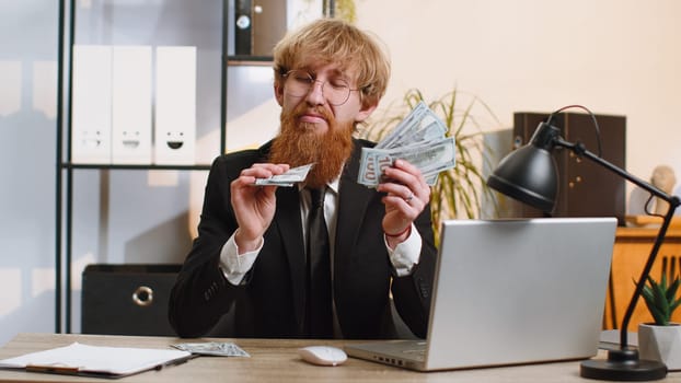 Rich pleased boss businessman guy waving money dollar cash banknotes bills like a fan success business career lottery winner, big income, wealth. Young bearded man freelancer at home office workplace