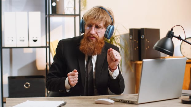 Happy relaxed overjoyed young businessman working on laptop computer at home office wearing headphones listening favorite energetic disco music on smartphone. Freelancer man relaxing, taking a break