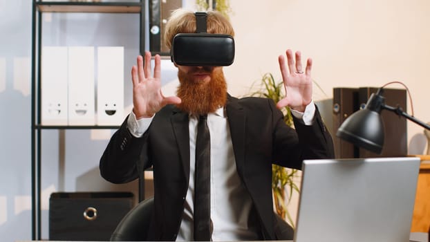 Young bearded businessman programmer software developer using headset helmet app to play simulation game. Watching virtual reality 3D 360 video. Young redhead in VR goggles working at home office