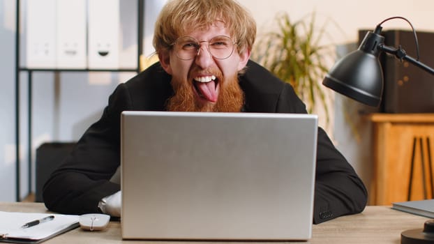 Redhead businessman hiding behind laptop computer, making funny face, fooling around, putting his tongue out remote working disrespecting someone at office desk workplace. Professional freelancer man