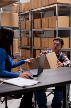 Warehouse manager taking parcel from worker in wheelchair to create invoice on laptop. Post office storehouse employees managing package distribution in disability friendly workplace