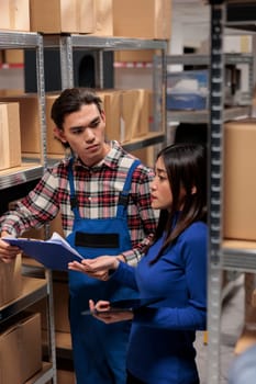 Asian man and woman doing warehouse logistics management together. Storehouse employees collaborating while analyzing inventory tracking and comparing data on digital tablet and clipboard
