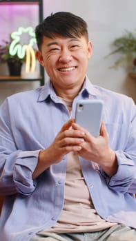 Asian man sits on sofa uses smartphone smiles at home room apartment. Chinese guy texting share messages content on mobile phone social media applications online, watching relax movie. Vertical shot