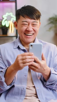 Happy asian winner man use mobile smartphone typing browsing say Wow yes found out great big win good news celebrate lottery jackpot. Chinese adult guy at home in room sitting on sofa. Vertical shot