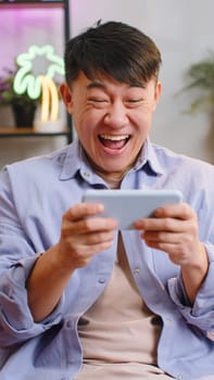 Worried asian winner man playing racing or shooter video games on smartphone at home. Chinese guy using smartphone gadget app with drive simulator sitting on couch in living room. Vertical shot