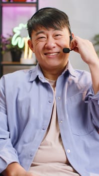 Asian handsome man wearing headset, freelance worker, call center or support service operator helpline, having talk with client or colleague communication support waving hello at home. Vertical