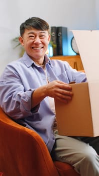 Happy asian man unpacking delivery parcel at home. Smiling satisfied chinese guy shopper online shop customer opening cardboard box receiving purchase gift by fast postal shipping. Vertical shot