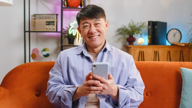 Asian man sitting on sofa uses mobile phone smiles at home living room apartment. Chinese adult guy texting share messages content on smartphone social media applications online, watching relax movie