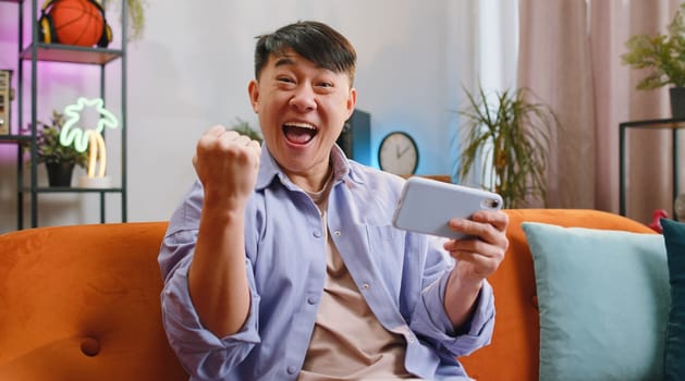 Worried asian handsome man enthusiastically playing racing or shooter video games on smartphone at home. Chinese guy using smartphone gadget app with drive simulator sitting on sofa in living room