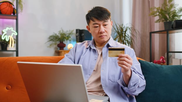 Asian man using credit bank card and laptop computer while transferring money, purchases online shopping, order food delivery at home apartment. Adult guy in day living room sitting on couch indoors