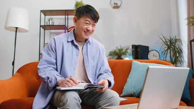 Online teacher e-learning in remote chat. Distance education online. Asian man at home making conference video call talking to webcam. Chinese student guy making notes on notebook sitting on sofa