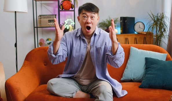 Happy asian man shouting wow, celebrating success, winning, goal achievement good news raising hands in gesture I did it, lottery luck at home apartment. Chinese guy in living room sitting on couch