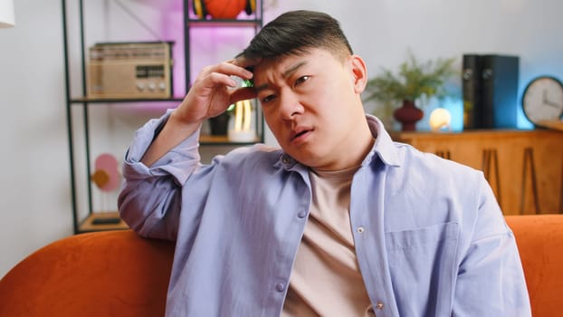 Sad lonely man sitting at home living room looks pensive thinks over life concerns, unrequited love suffers from unfair situation. Chinese guy problem, break up, depressed feeling bad annoyed burnout