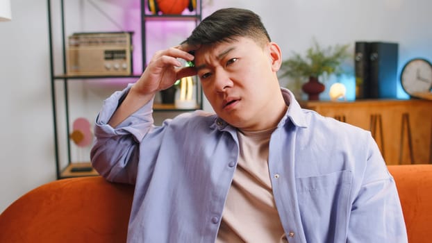 Sad lonely man sitting at home living room looks pensive thinks over life concerns, unrequited love suffers from unfair situation. Chinese guy problem, break up, depressed feeling bad annoyed burnout