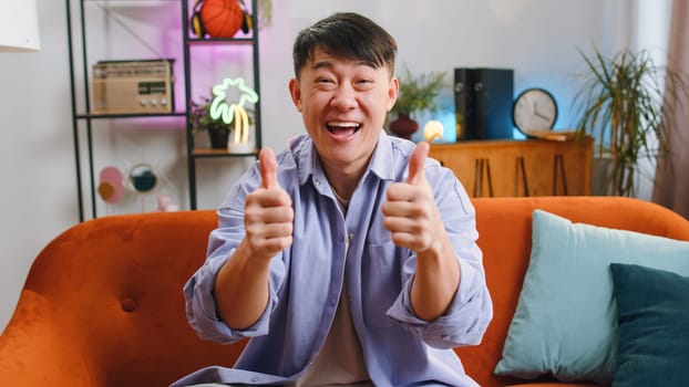 Like. Happy excited asian man looking approvingly at camera showing thumbs up, like sign positive something, good great news, positive feedback. Adult chinese guy sitting on couch at home living room