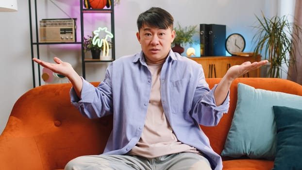 I dont know what to say. Confused asian handsome man feeling embarrassed about ambiguous question, having doubts, no idea, being clueless and uncertain at home. Chinese guy sits on sofa in living room