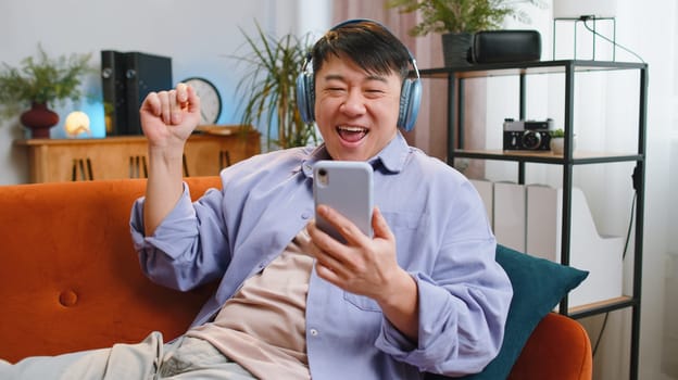 Happy overjoyed asian man in wireless headphones relaxing sitting on couch at home apartment choosing listening favorite energetic disco dancing music. People weekend daytime leisure activities