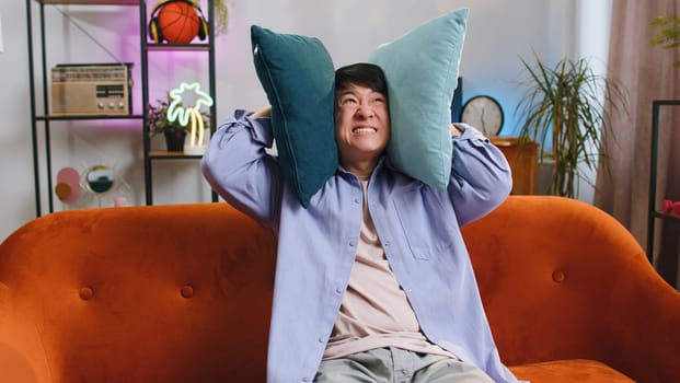 Repair work at neighbours. Irritated asian man relaxing on couch cover ears with pillows annoyed by noisy neighbors suffer from headache wish silence. Thin walls at home flat without sound insulation