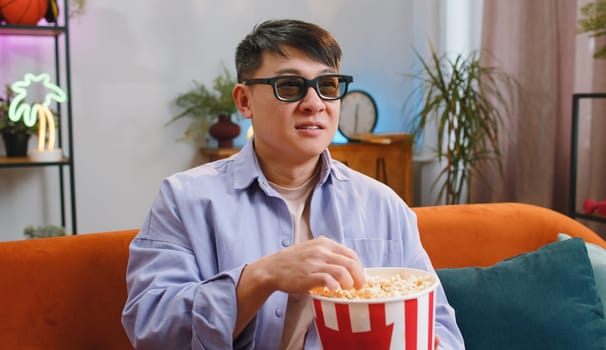 Excited asian man in 3D glasses sits on couch eating popcorn snacks and watching interesting TV serial sport game, film online social media movie content at home apartment. Guy enjoying entertainment