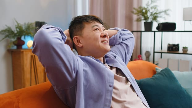 Portrait of tired asian man enjoy relaxing on home couch in room. Happy chinese guy lying on couch resting napping after hard working day, closed his eyes, put his hands behind his head, stress free